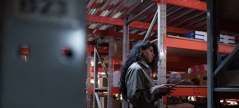 A woman in a uniform, looking in an AC storage unit at shelves with a checklist in her hands;